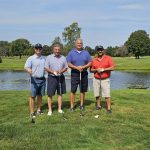 Four men stand on a grassy golf course near a pond, each holding a golf club. The sky is clear with a few clouds, and trees line the background. They are smiling and dressed in casual golf attire.