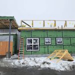 A person stands near a partially constructed building covered in green weatherproof sheathing. Snow covers the ground, and construction materials, ladders, and a dumpster are visible around the site.