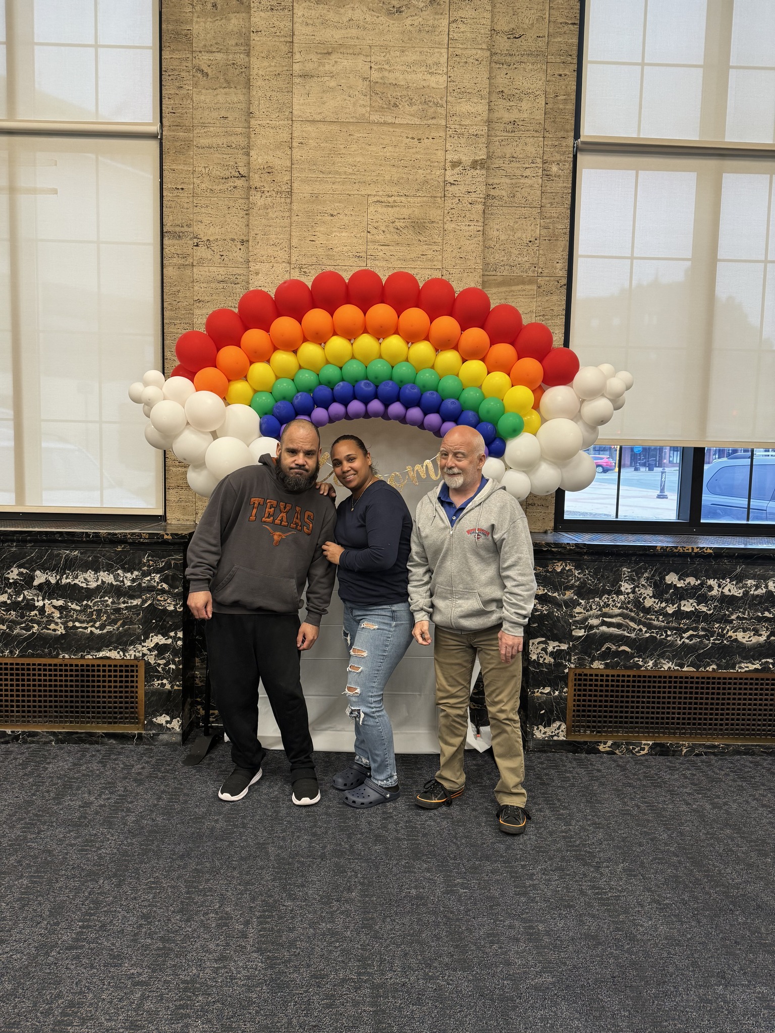 Three people stand smiling in front of a colorful rainbow balloon arch with white balloons as clouds in a room with tall windows and dark carpet.