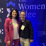 Two women stand smiling in front of a navy blue backdrop that reads The Womens Edge and Mentor. Partner. Lead. Both are wearing business attire with name tags.