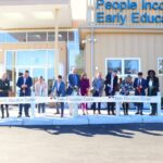 A group of people stand in front of an Early Education Center, holding large scissors and cutting a ribbon at a grand opening ceremony on a sunny day.