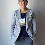 A person with short hair, glasses, and earrings smiles while wearing a light gray blazer, navy shirt, floral boutonniere, and a name tag with ribbon badges against a light textured wall.