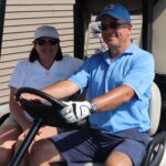 A smiling man and woman sit in a golf cart. The man, wearing a blue shirt, cap, and sunglasses, is driving. The woman, in a white shirt, hat, and sunglasses, sits beside him. Golf clubs are visible in the background.