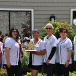 Six women wearing matching white shirts pose together outdoors, smiling. One woman in the center holds a decorative gold star. Green plants, a window, and a tent are visible in the background.