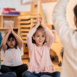 Three young children sit cross-legged on the floor with their arms raised above their heads, mimicking a teacher who is leading an activity in a classroom setting. The atmosphere appears positive and engaging.
