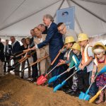 A group of adults in suits and children in hard hats use colorful shovels to break ground at a ceremony under a white tent, smiling and lined up along a mound of dirt.