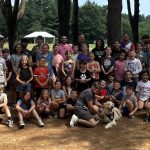A large group of children and adults pose together outdoors in a wooded park area, some wearing yellow shirts. A dog sits at the front, and picnic shelters are visible in the background.