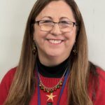 A woman with long brown hair, glasses, and a red sweater featuring a snowman design smiles at the camera. She is wearing festive jewelry and a lanyard, with a plain light-colored background behind her.