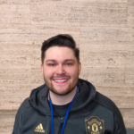 A man with short dark hair and a beard smiles at the camera. He is wearing a dark Manchester United jacket, a blue lanyard, and is seated in front of a beige stone wall.