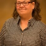 A woman with curly brown hair, glasses, and hoop earrings, wearing a black and white houndstooth patterned shirt, stands in front of a plain brown background, smiling slightly.