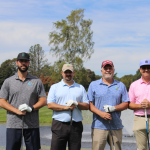 Four men stand side by side on a golf course, all holding golf clubs and wearing polo shirts, caps, and sunglasses. A fountain and trees are visible in the background under a partly cloudy sky.