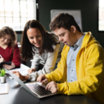Three young adults work together in a modern office. One man in a yellow jacket types on a laptop while a woman leans in to help. Another woman sits in the background, writing in a notebook.