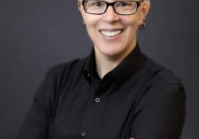 A person with short, shaved hair on the sides and longer hair styled upward, wearing glasses and a black button-up shirt, smiles with arms crossed against a dark, plain background.