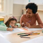 A young girl draws with colored markers on paper while a smiling adult sits beside her, watching and holding a pen. Art supplies are spread on the table in a bright classroom.